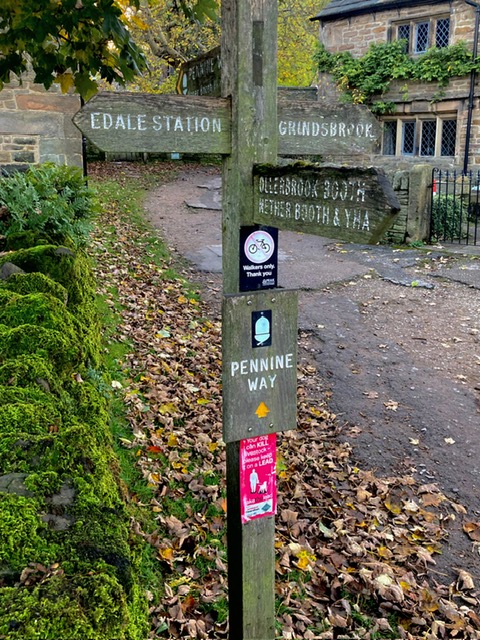 Wooden signpost with directions to Edale Station and Pennine Way, surrounded by lush greenery and rustic stone buildings