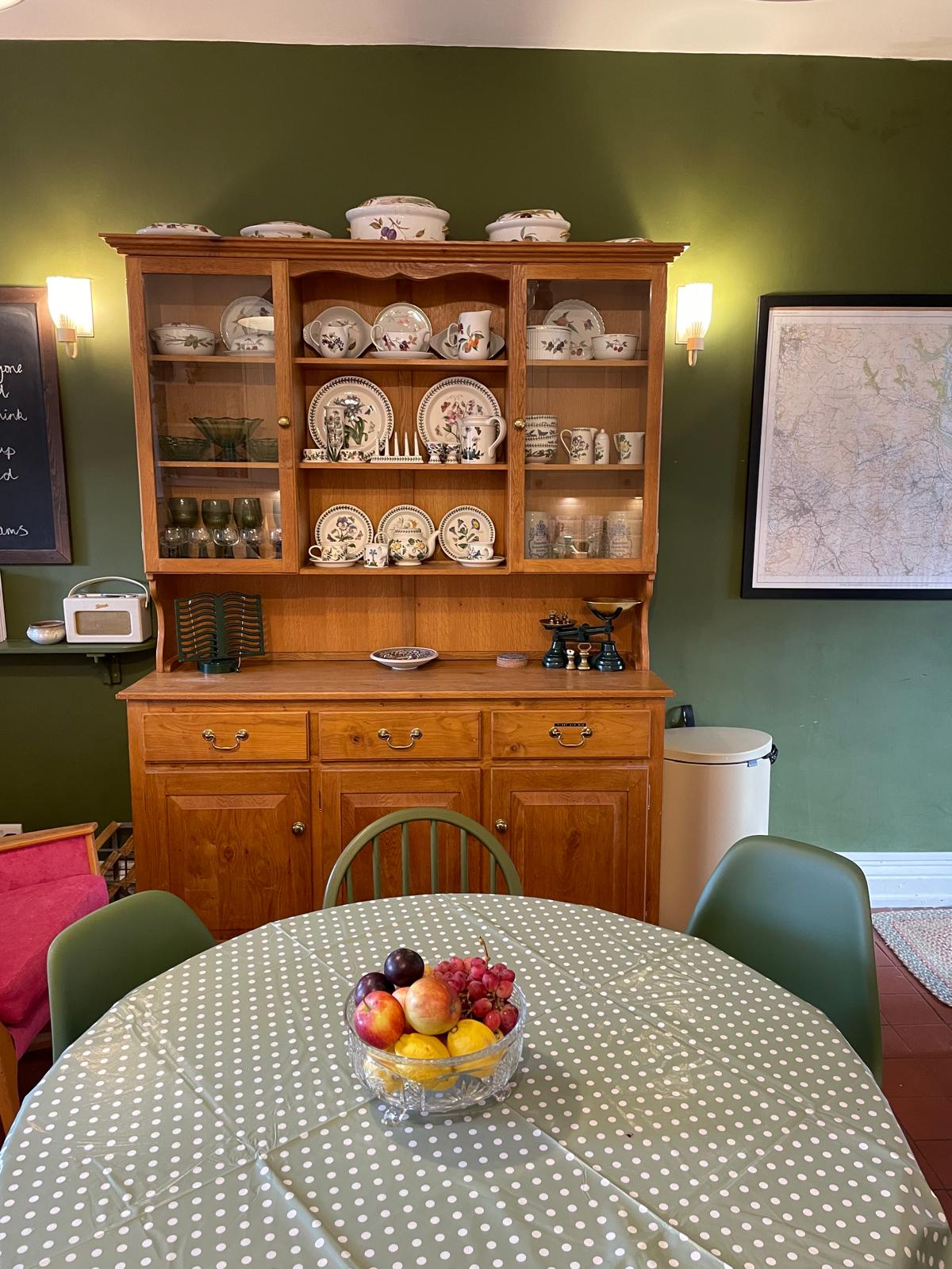 Charming cottage kitchen with wooden hutch, green walls, round table, and fresh fruit centerpiece. Cozy and inviting atmosphere