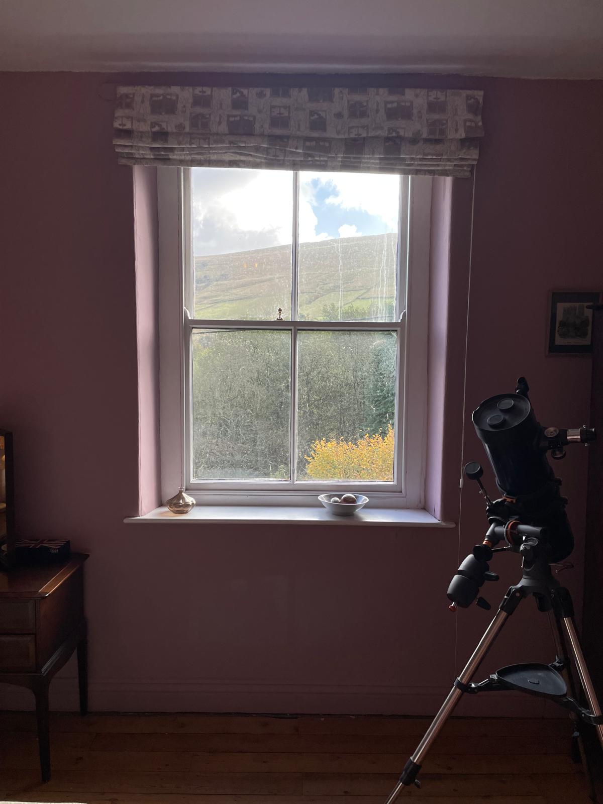 Cozy bedroom with a telescope, pink walls, wooden floor, and a scenic view of the mountains through a large window