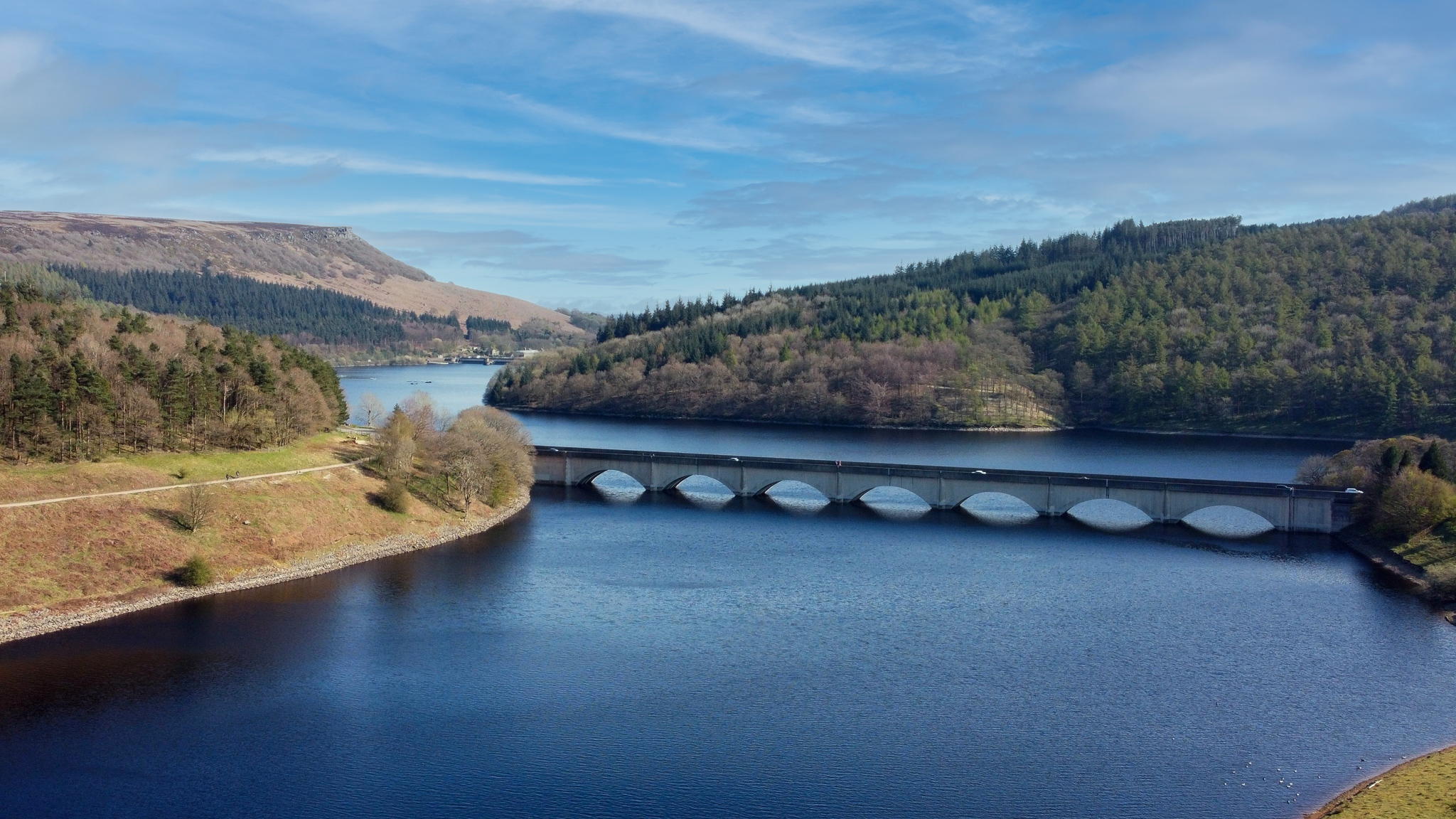 Scenic view of a serene lake with a modern bridge, surrounded by lush green hills and blue skies